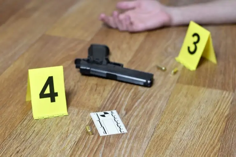 A picture of a person's arm on the floor that is dead from a firearm. A gun is on the floor nearby.